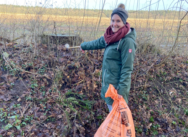 Junge Frau sammelt Müll aus der Landschaft und hat einen orangenen AWD Sack in der HAnd.