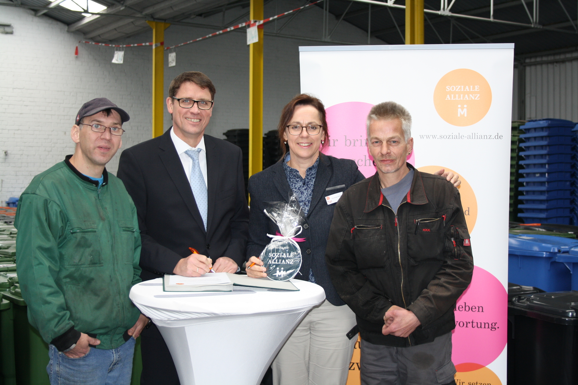 Vier Personen der Sozialen Allianz stehen vor einem Werbebanner mit dem Logo der Sozialen Allianz.