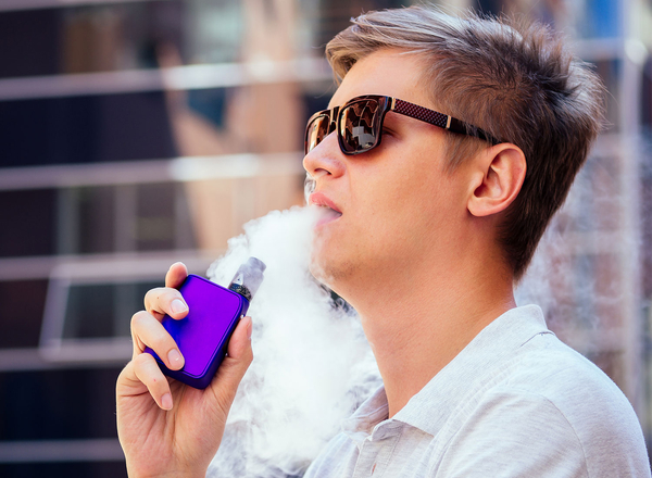 Junger Mann mit Sonnenbrille zeiht an einer Vape, so dass Qualm Dampf sichtbar ist.